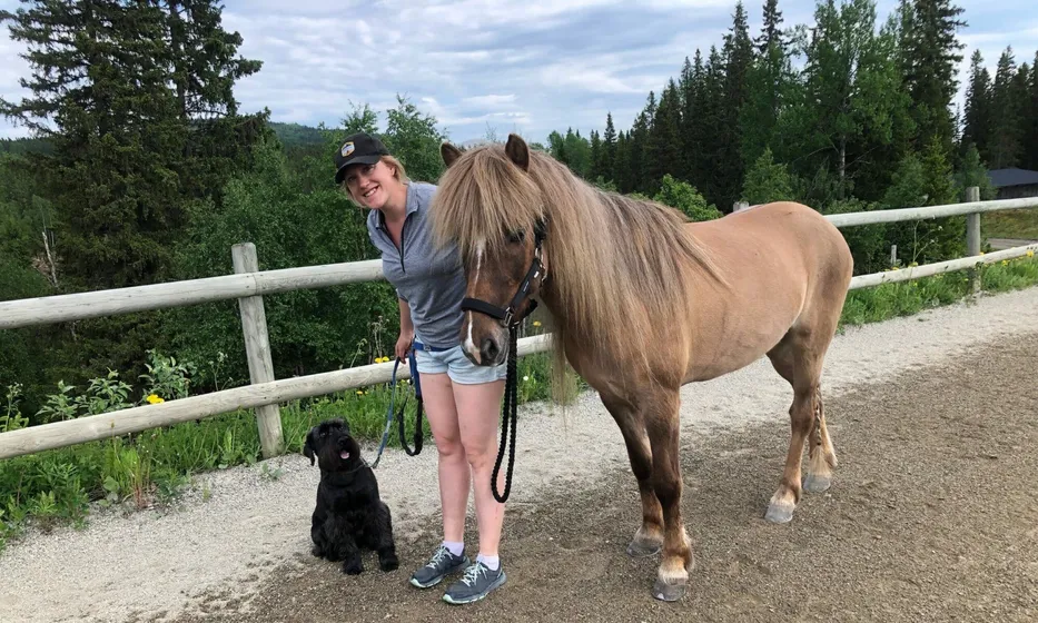 Malin Stang står bredvid en brun häst. Bredvid sitter en svart hund. Sommarbild.